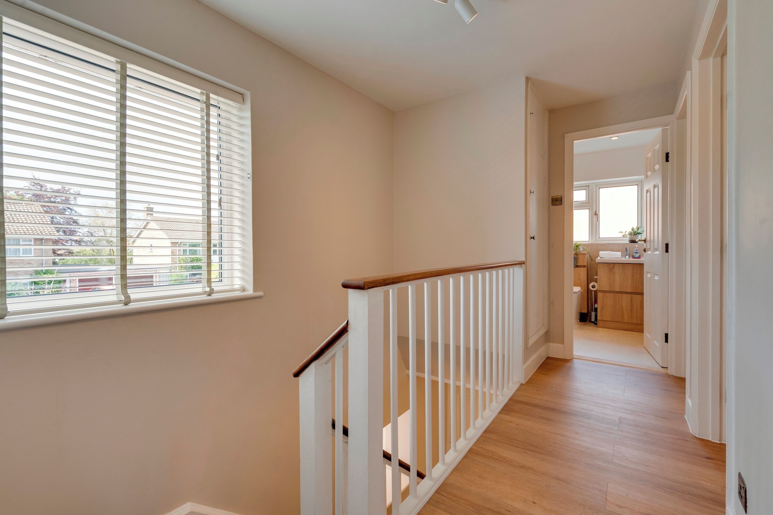 A hallway with a wooden floor, white railing, and a window with blinds.