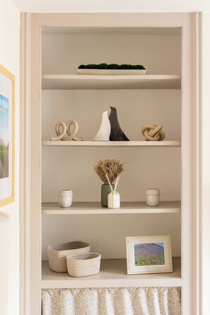 Une étagère blanche ornée d’objets décoratifs, notamment un bol, un vase avec des fleurs séchées et un tableau encadré.