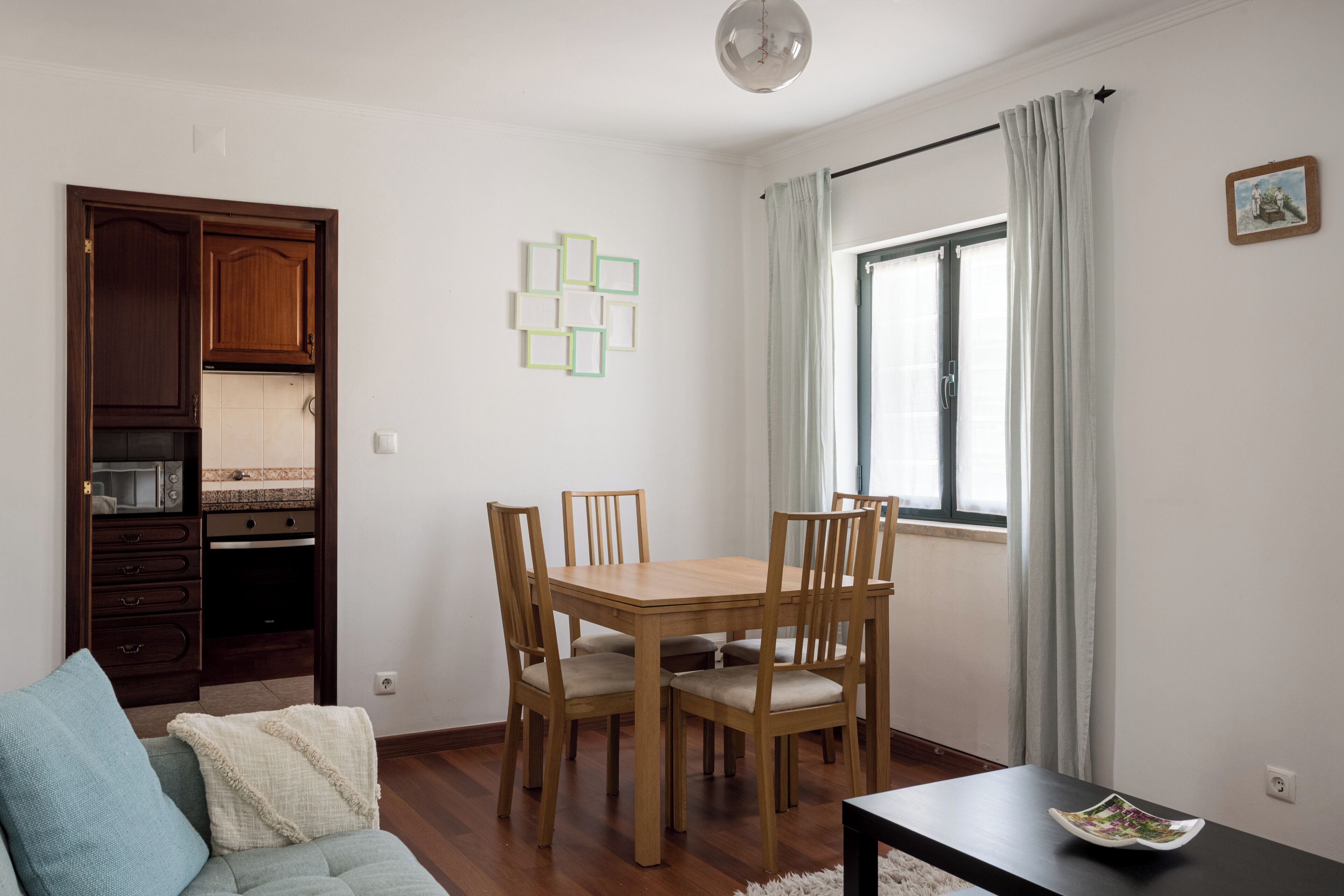 A dining area with a wooden table and chairs, a kitchen with cabinets, and a window with curtains.
