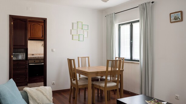 A dining area with a wooden table and chairs, a kitchen with cabinets, and a window with curtains. - Lisbon Olivais Residence – 2BR Apartment (Lisbon)