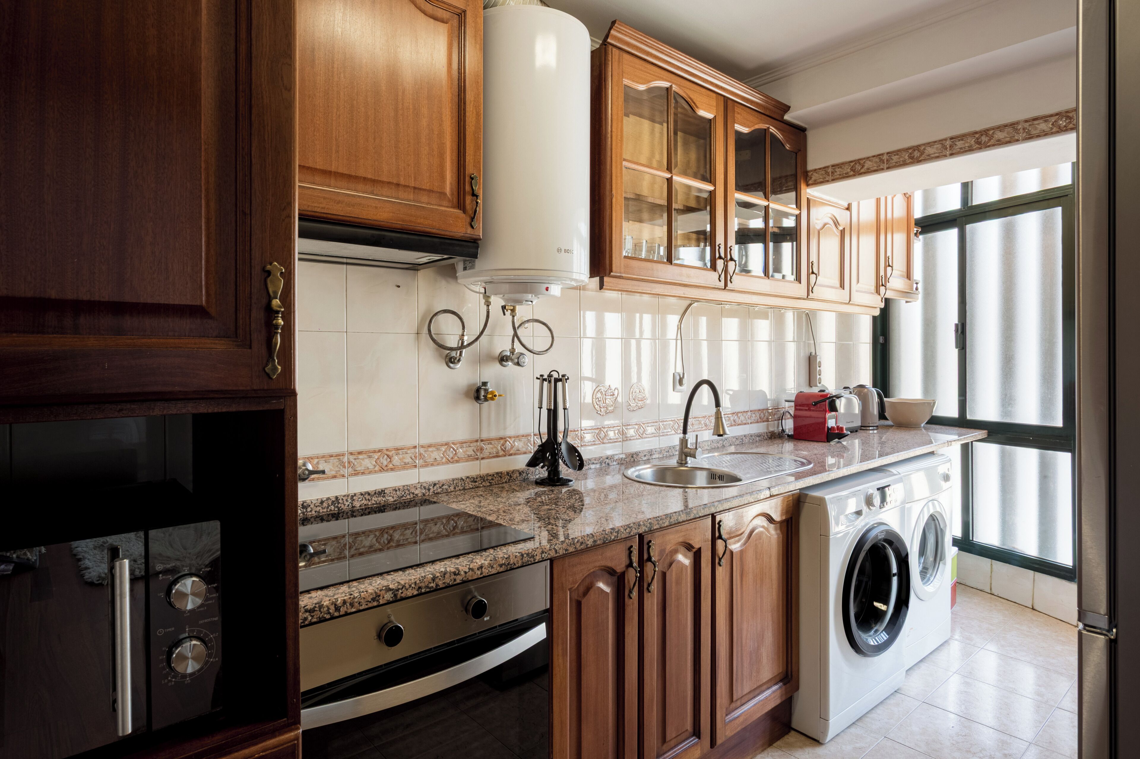 A modern kitchen with wooden cabinets, a built-in oven, a washing machine, and a window providing natural light.