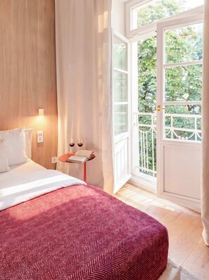 A hotel room with a bed, a bedside table with two wine glasses, a book, and a view of greenery through a large window. - TurnKey | Elegant Studio for 2 | Republic Square (Hlavní město Praha)