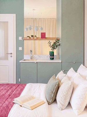 A bedroom with a bed, pillows, a bedside table with a book, a mirror, and a small kitchenette area. - TurnKey | Elegant Studio for 2 | Republic Square (Hlavní město Praha)