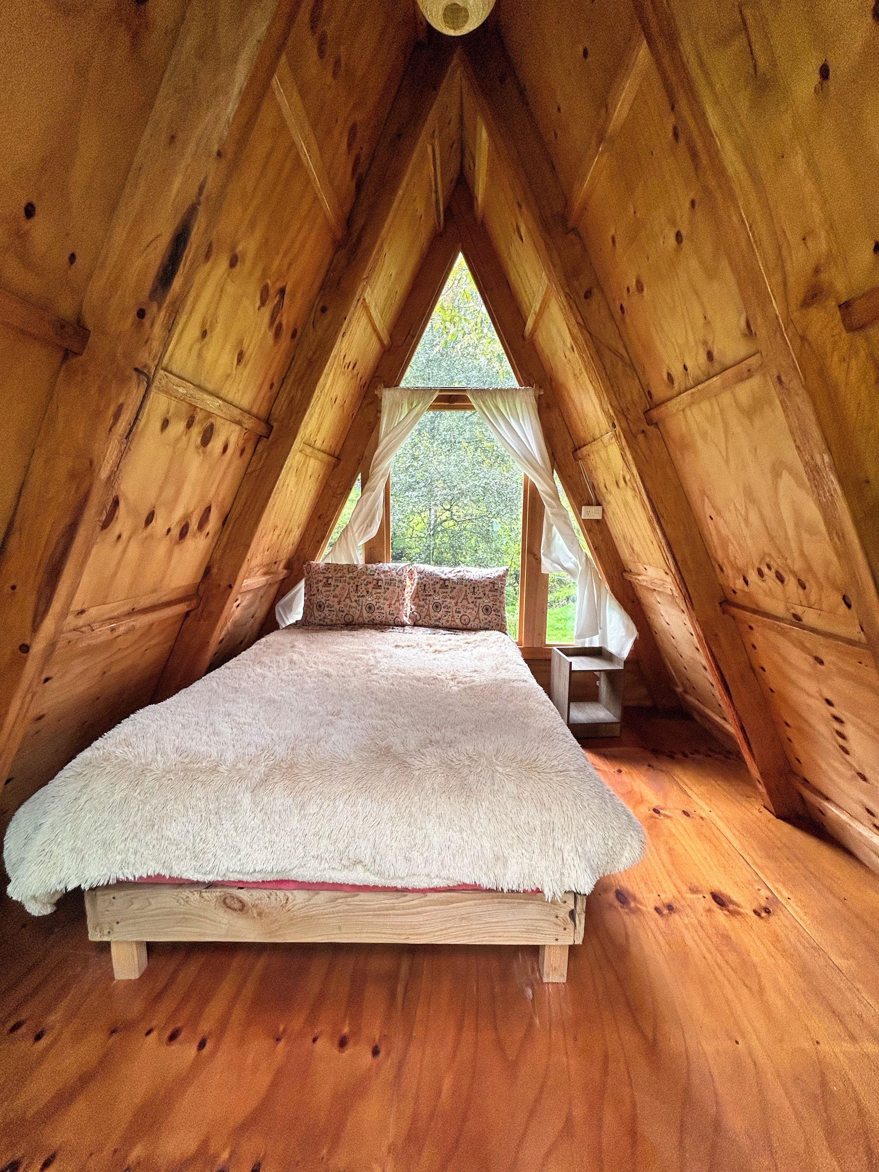 Un dormitorio acogedor en el ático con techo inclinado de madera, una cama con edredón blanco y una ventana con cortinas.
