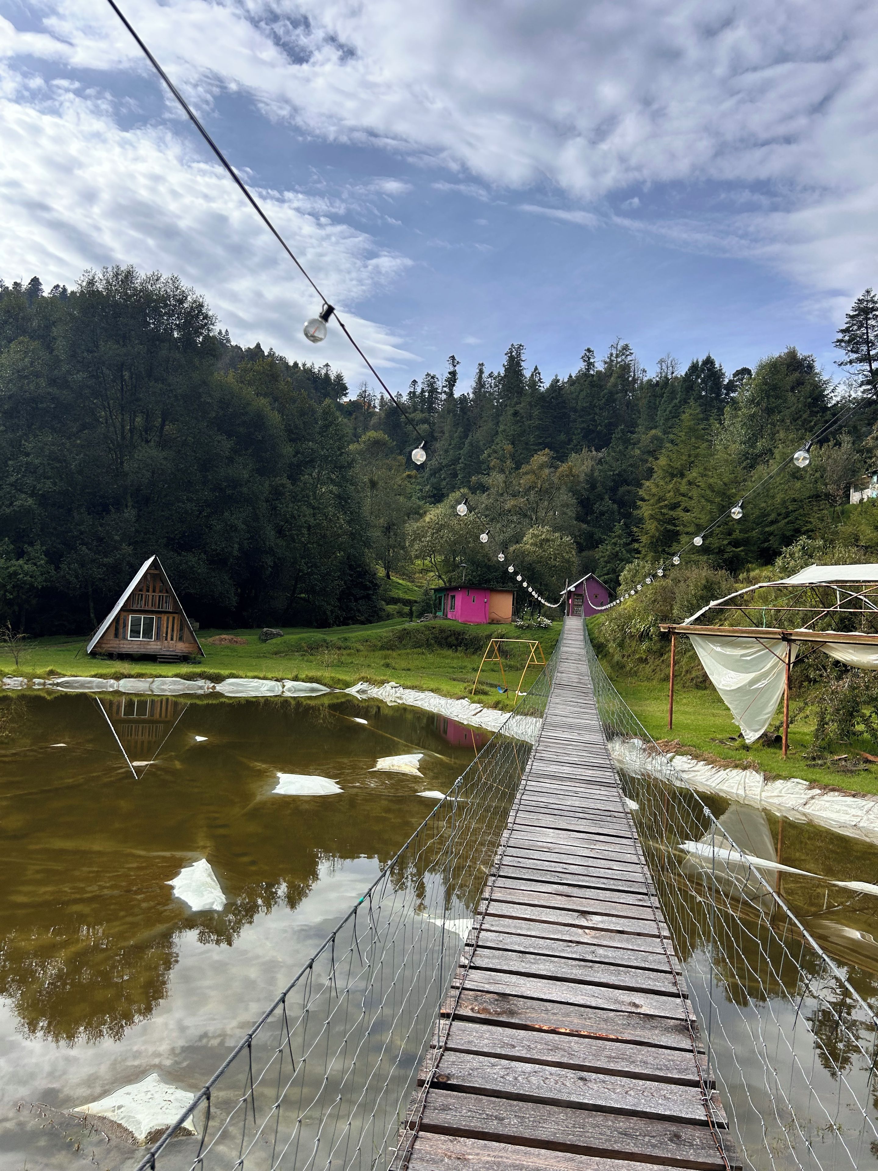 Vesialueen yllä kulkeva riippusilta, jossa on puinen kävelytaso. Sillan ympärillä on metsäisiä kukkuloita ja lähellä sijaitsee pieni mökki.