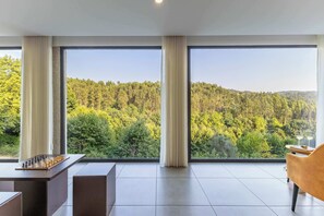A modern room with large windows, a chessboard, and a view of a forested hillside.