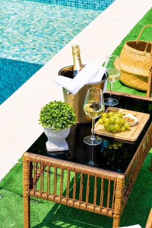 A poolside setting with a table featuring a bottle of champagne, a glass of white wine, a bowl of grapes, and a small potted plant.