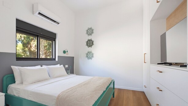 Een slaapkamer met een bed, een raam, een airconditioner, een wanddecoratie en een dressoir.