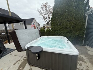 A hot tub with a black cover, a wooden deck, and a tall evergreen tree in the background. - Couples Escape w/ Hot Tub & Fire Pit Near the Lake (Clarklake)