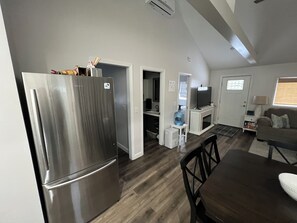 A modern kitchen with a stainless steel refrigerator, a dining area with a dark wooden table, and a living room with a sofa and a TV. - Couples Escape w/ Hot Tub & Fire Pit Near the Lake (Clarklake)