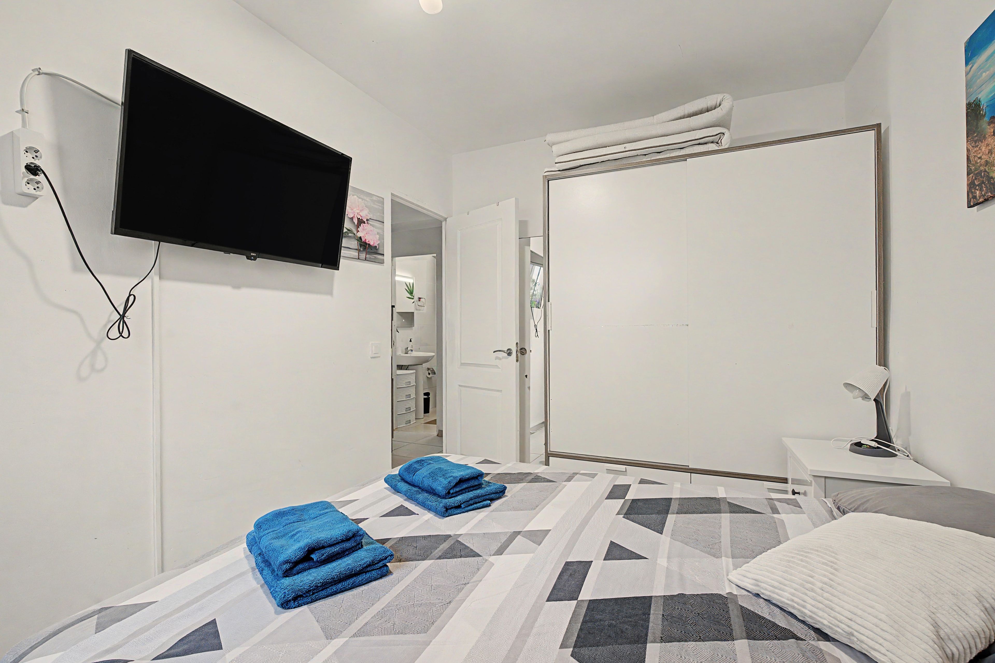 Habitación de hotel con cama, televisión montada en la pared, mesita de noche con lámpara y baño a la vista a través de una puerta abierta.