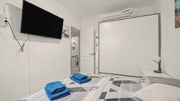Habitación de hotel con cama, televisión montada en la pared, mesita de noche con lámpara y baño a la vista a través de una puerta abierta.