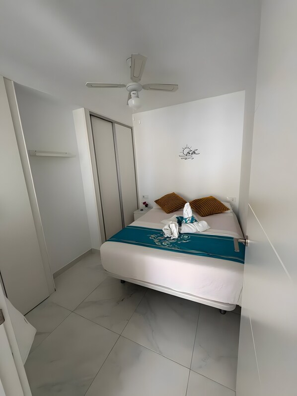 A hotel room with a bed, a ceiling fan, a wall-mounted light, and a door.