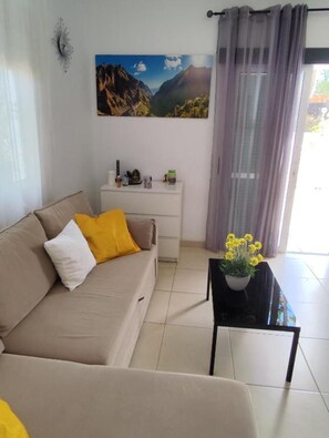 A living room with a beige sofa, a small white nightstand, a black coffee table with a potted plant, and a painting of a mountain landscape on the wall.