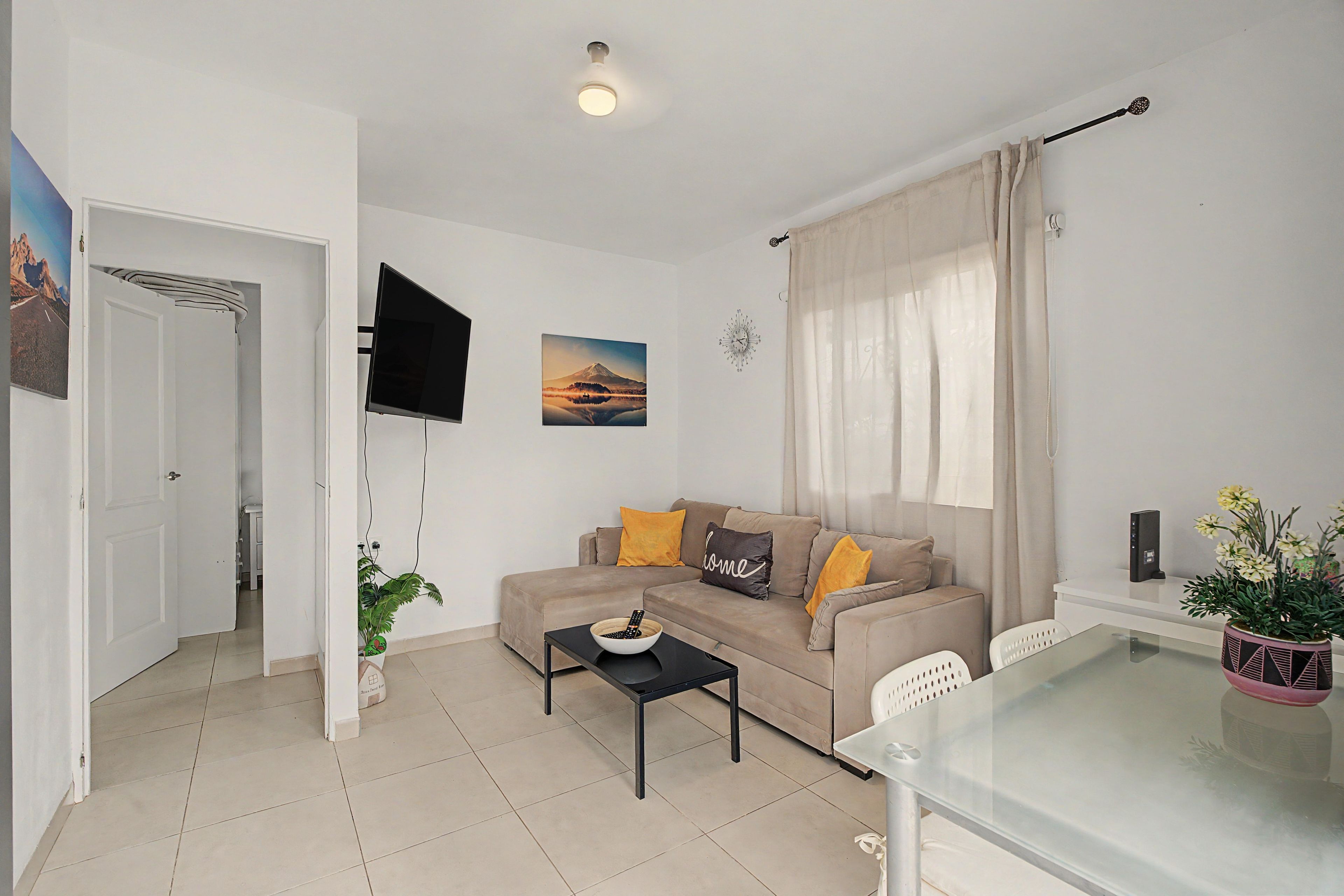 A modern living room with a sofa, a coffee table, a TV mounted on the wall, and a dining table with chairs.