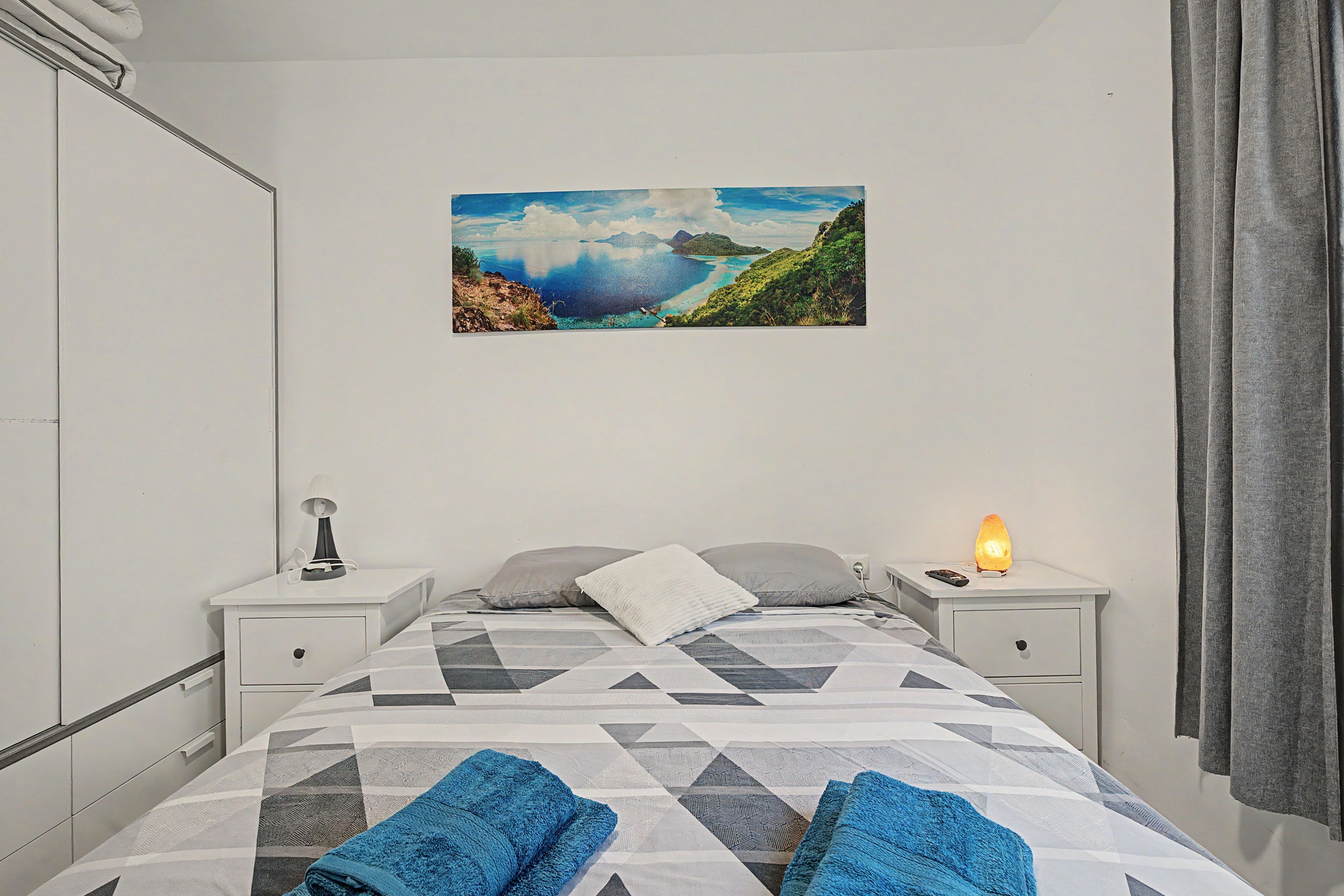 A bedroom with a bed, two bedside tables, a night lamp, a heater, and a painting of a landscape.