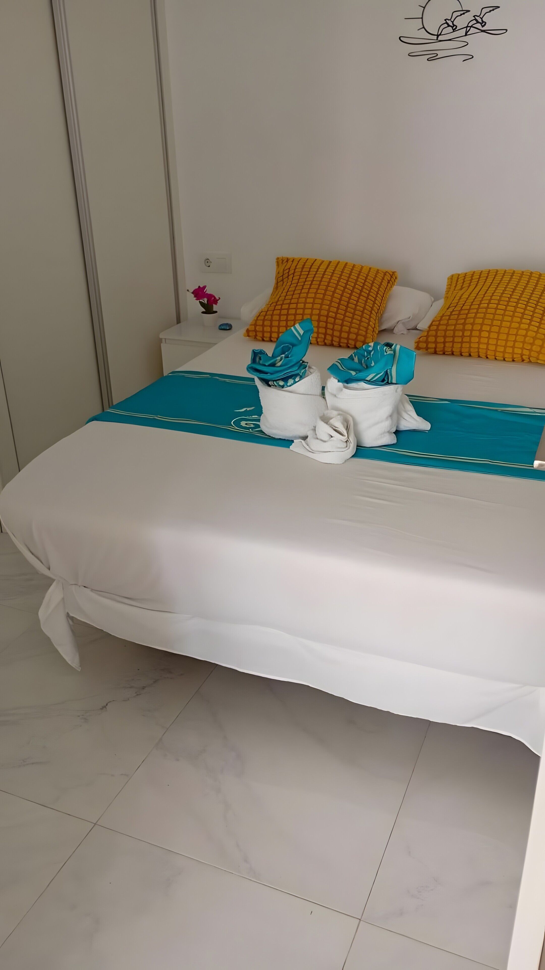 Una cama bien hecha con ropa blanca y toallas azules, una almohada amarilla y una pequeña planta en maceta sobre la mesita de noche.