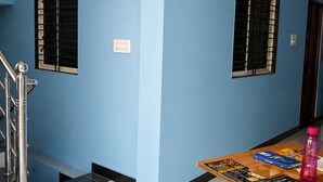 Um quarto com parede azul, uma escada, uma mesa com livros e uma garrafa rosa, e uma janela com persiana preta.