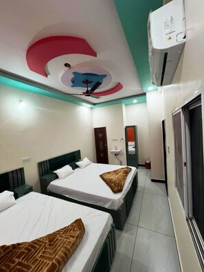 A hotel room with two beds, a ceiling fan, an air conditioning unit, and a bathroom with a mirror and sink. - Hotel Kridhika Inn (Ujjain)