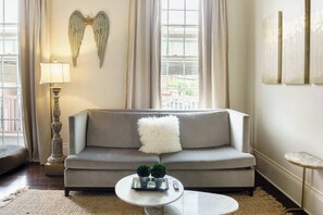 A living room with a grey sofa, a white fluffy pillow, a round coffee table with a tray and two potted plants, a lamp, and a wall-mounted angel decoration. - LaBranche Pharmacy | Step Back in Time in a Historic Tremé Landmark- Walk to French Quarter (New Orleans)
