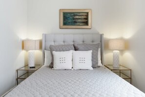 Una cama bien tendida con cubrecama acolchado, dos lámparas de noche y una obra de arte enmarcada sobre el cabecero.