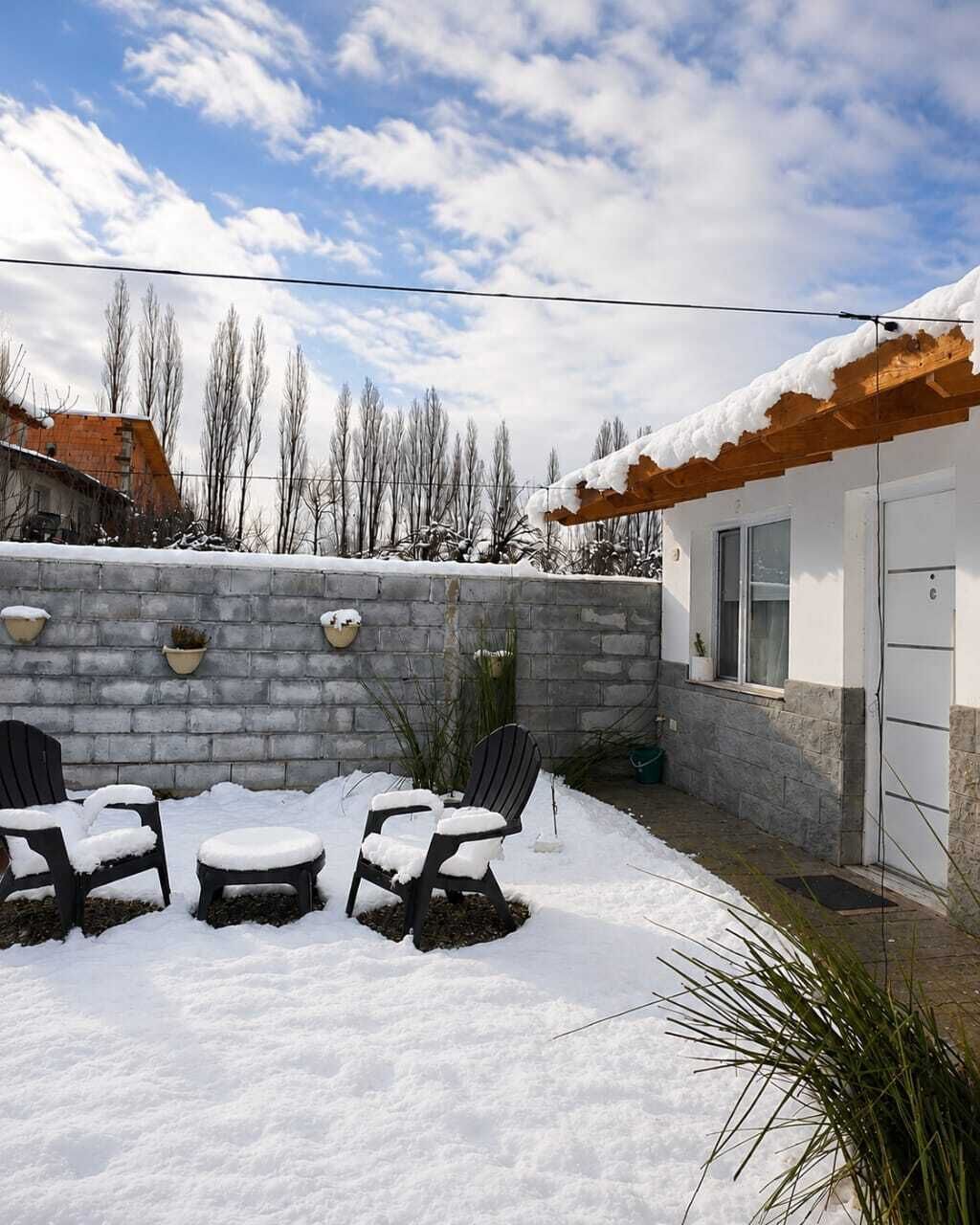 Um pátio com três cadeiras cobertas de neve, um muro de pedra e um edifício com telhado de madeira.