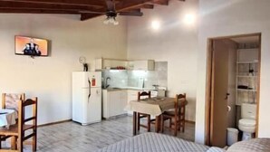 Une pièce avec un plafond en bois, un lit recouvert d’une courtepointe, un bureau avec une chaise et, au fond, un coin cuisine.