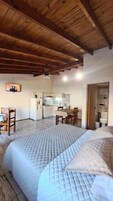Una habitación con techo de madera, cama con cubrecama acolchado, escritorio con silla y una zona de cocina al fondo.