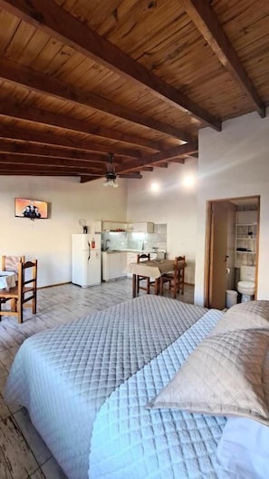 Une chambre avec un grand lit, des poutres apparentes en bois au plafond, un petit bureau et, au fond, une kitchenette.