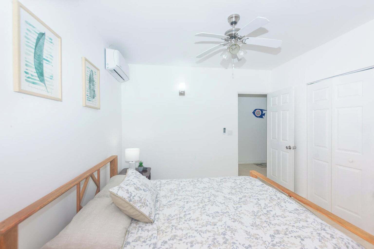 A bedroom with a bed, a ceiling fan, a wall-mounted air conditioner, and a door leading to another room.