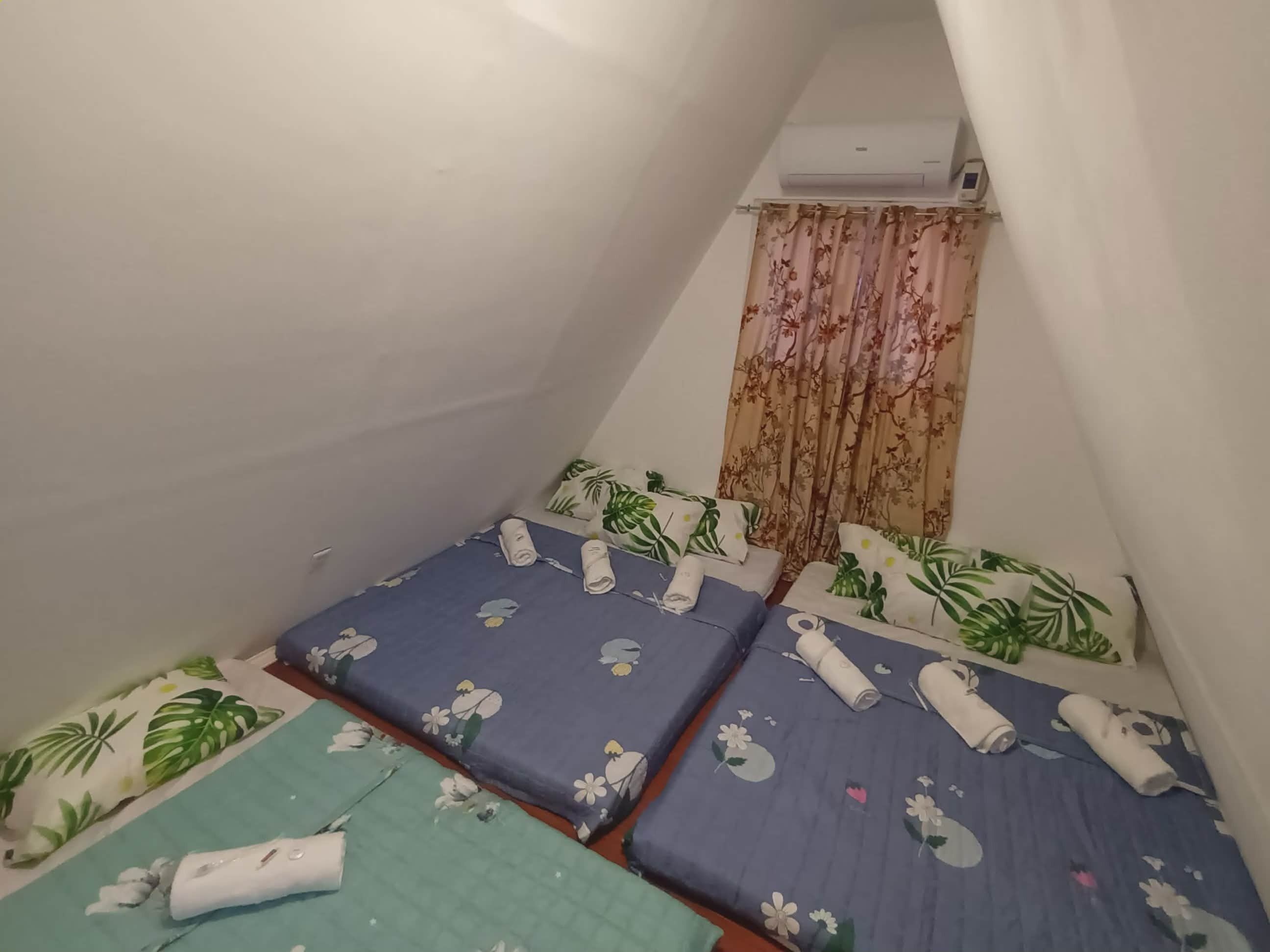 Une petite chambre compacte avec trois lits, chacun recouvert d’une courtepointe à motifs floraux et agrémenté de serviettes enroulées. Un appareil de climatisation est fixé au mur, au-dessus des lits.