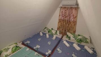 Une petite chambre compacte avec trois lits, chacun recouvert d’une courtepointe à motifs floraux et agrémenté de serviettes enroulées. Un appareil de climatisation est fixé au mur, au-dessus des lits.