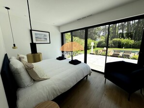 A modern bedroom with a large bed, a side table, a lamp, and a view of a garden through a sliding door. - Villa Murmur - Seignosse - Pool - Jacuzzi (Seignosse)