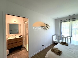 Ein Schlafzimmer mit einem großen Bett, einem Fenster mit Vorhängen, einem weißen Babybett und einem Badezimmer mit Spiegel und Waschbecken.