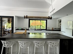 A modern kitchen with a large island, white chairs, and a view of greenery outside. - Villa Murmur - Seignosse - Pool - Jacuzzi (Seignosse)