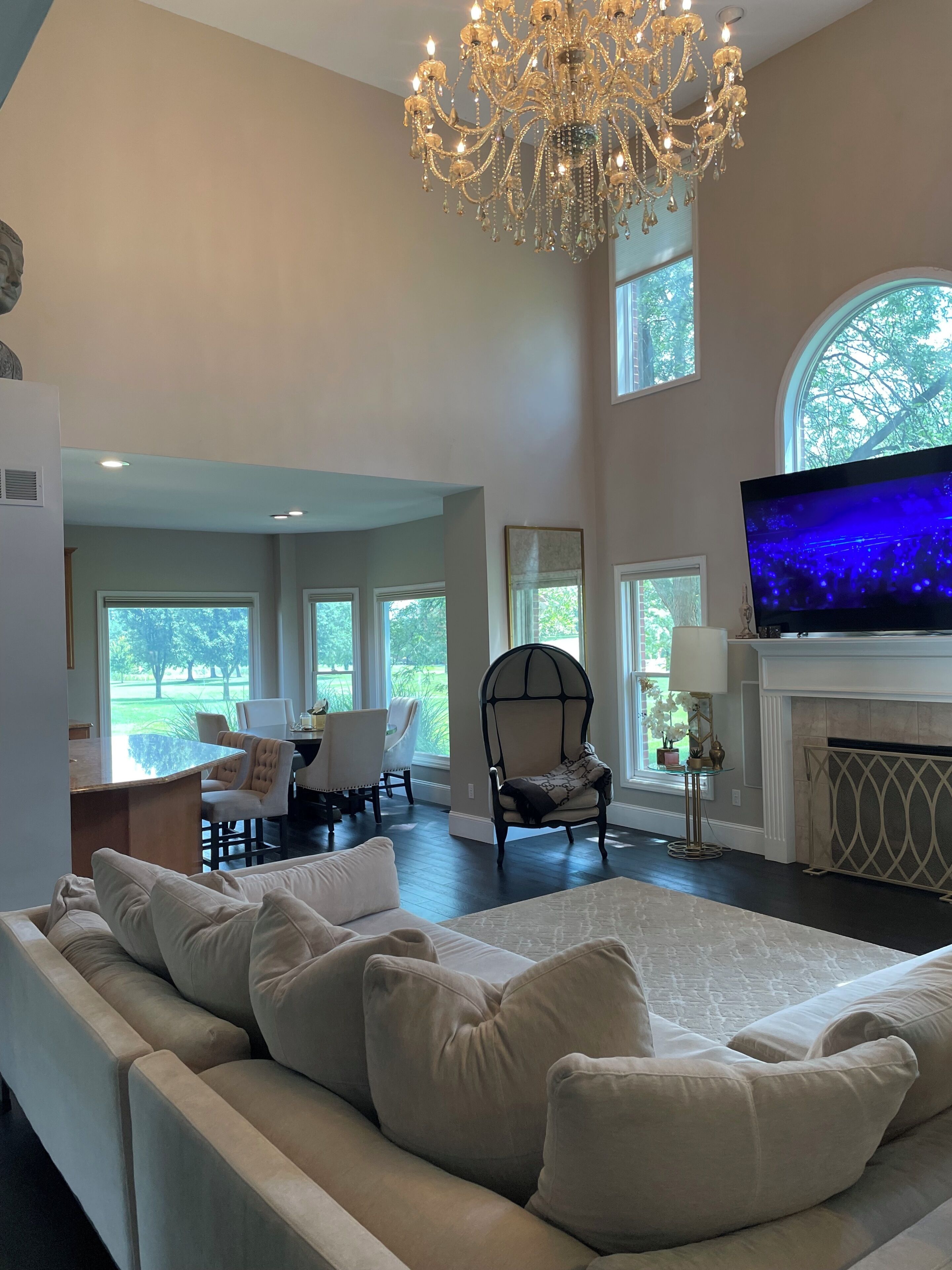 A spacious living room with a large sofa, a television, and a fireplace. There are multiple windows providing natural light and views of the outdoors.