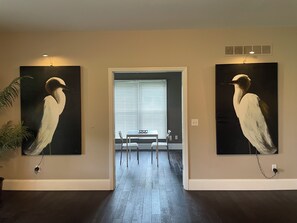 A hallway with two wall-mounted paintings of birds, a dining table with chairs, and a window with blinds.