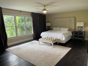 A bedroom with a large bed, a nightstand, a lamp, a ceiling fan, and a view of greenery outside.