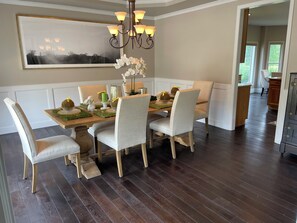 A dining area with a wooden table, white upholstered chairs, a chandelier, and a large framed picture on the wall.