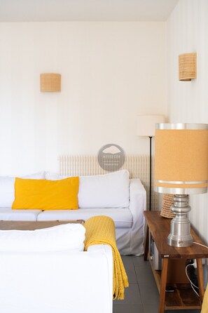 Una stanza con un divano bianco, una coperta gialla, una lampada su un comodino in legno e un termosifone.