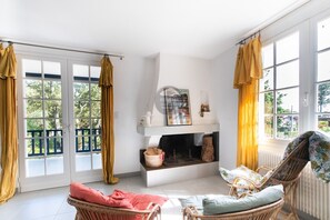 A bright room with a fireplace, wicker furniture, and large windows with curtains. - Villa Her Den - Hossegor - Town center - Lake (Soorts-Hossegor)