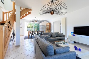 A modern living room with a sofa, a TV, and a dining area with chairs and a table. - Villa Fougères - Seignosse - 8 Adts+2 kids - Pool (Seignosse)