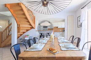 Une salle à manger avec une table en bois dressée pour six personnes, un escalier et une cuisine en arrière-plan.