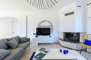 A modern living room with a grey sofa, a white coffee table with magazines, a fireplace, and a wall-mounted TV. - Villa Fougères - Seignosse - 8 Adts+2 kids - Pool (Seignosse)