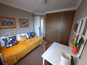 Ein Schlafzimmer mit einem Bett, einem weißen Tisch und einem Kleiderschrank. An der Wand hängen zwei Gemälde.