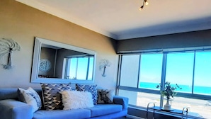A living room with a large window, a sofa, and a coffee table. - Amazing View For Less Bloubergstrand Beachfront (Cape Town)