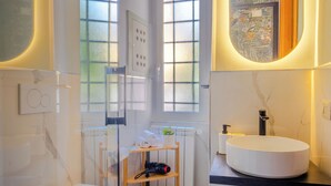 A modern bathroom with a white sink, a toilet, and a shower. There are two windows with frosted glass, a towel rack, and a shelf with toiletries.