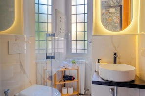 A modern bathroom with a white sink, a toilet, and a shower. There are two windows with frosted glass, a towel rack, and a shelf with toiletries.