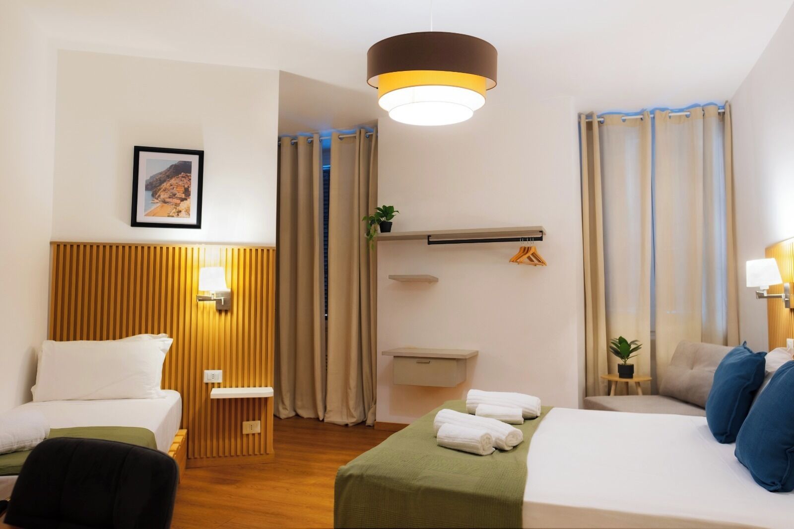 A hotel room with a bed, a desk, a chair, a bedside table, a lamp, a hanging plant, and a window with curtains.