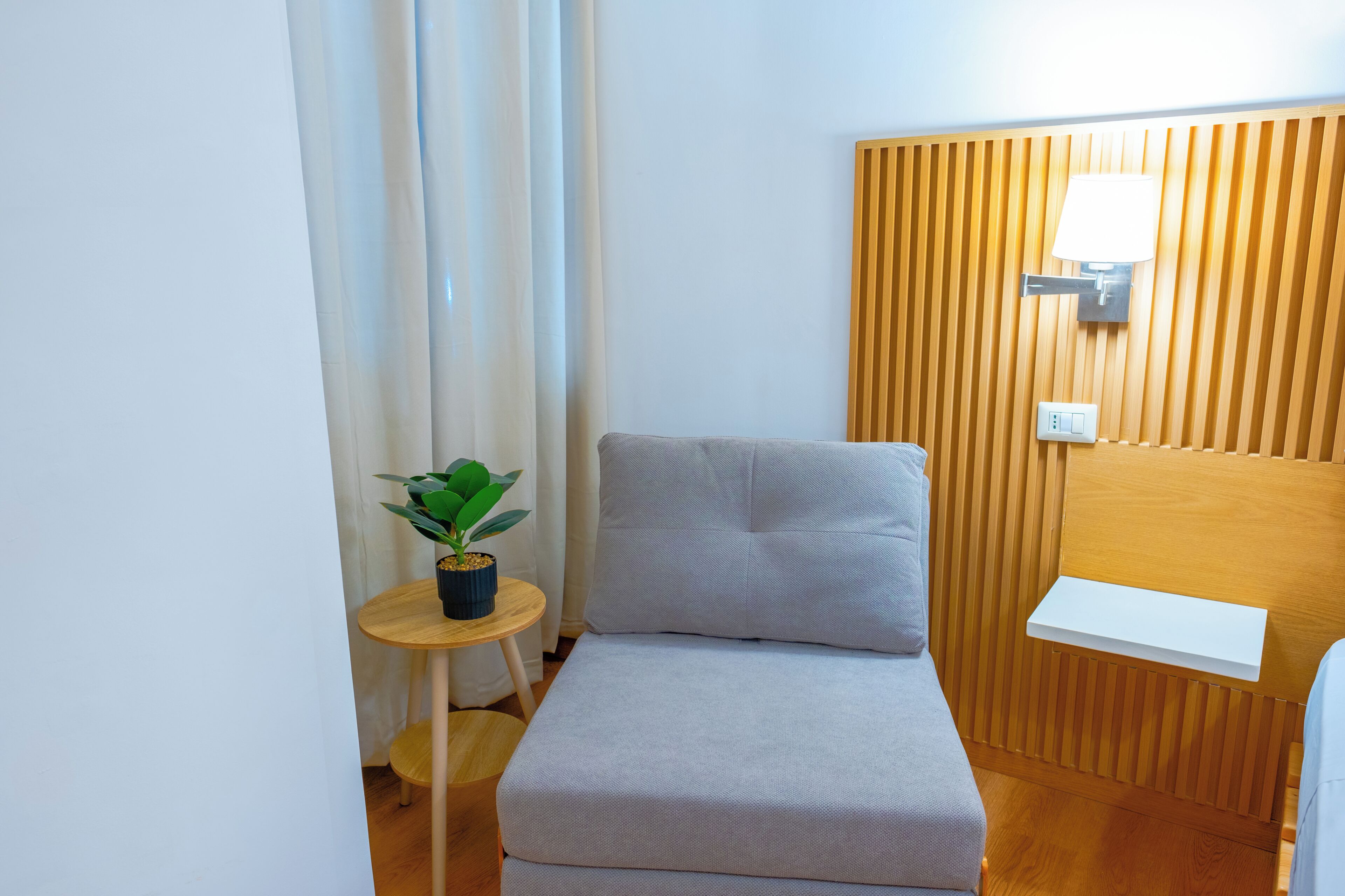 Een compacte, verzorgde kamer met een enkel bed, een nachtkastje met een plant, een wandlamp en een houten wandpaneel.